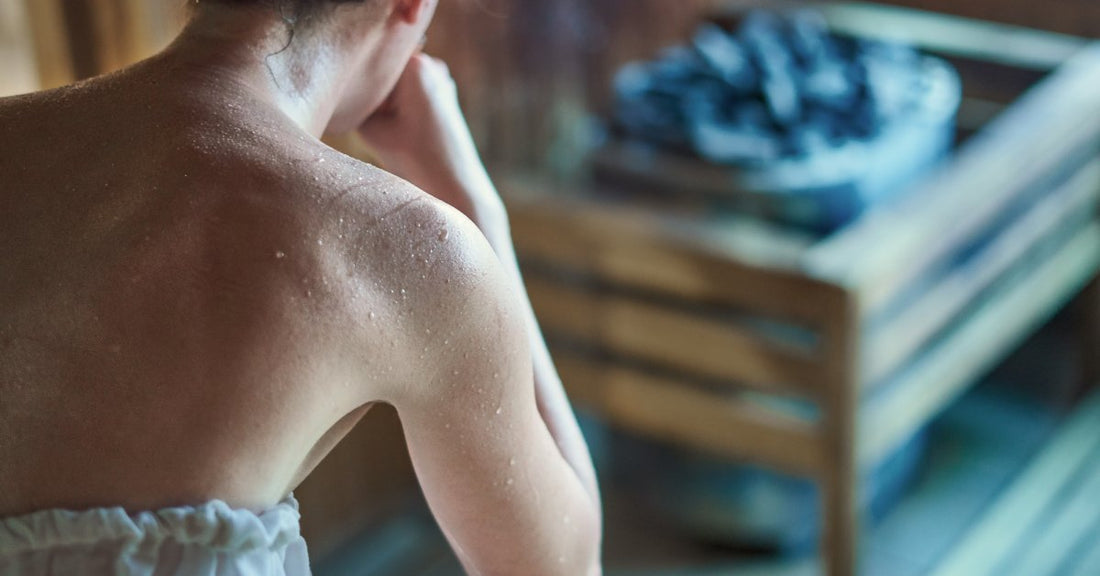 A woman wrapped in a white towel sits in front of a sauna stove filled with hot rocks. Her skin is noticeably sweaty.