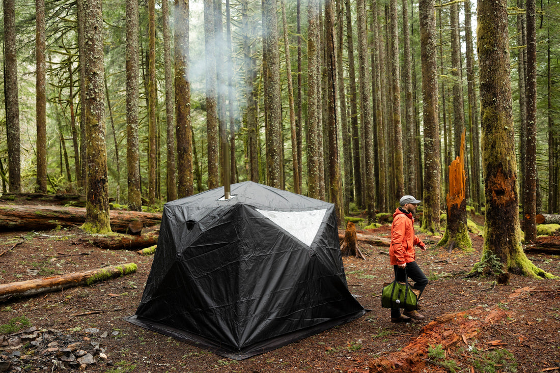 Portable wood-fired sauna tent set up outdoors on the shore of a lake, with smoke rising from the chimney.