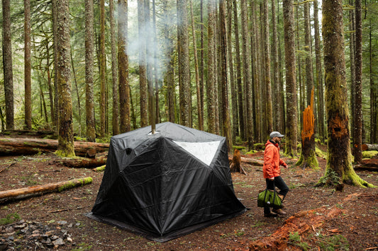 Portable wood-fired sauna tent set up outdoors on the shore of a lake, with smoke rising from the chimney.