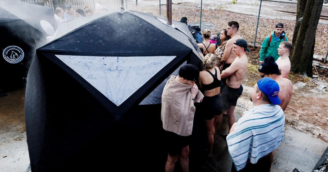 A highly-insulated, canvas portable sauna tent with a small wood-burning stove visible inside, set up outdoors near a lake or wooded area.