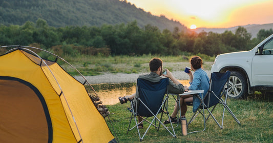 Two people sit in folding camp chairs and look at the sunset through lush green hills. A yellow tent sits nearby.