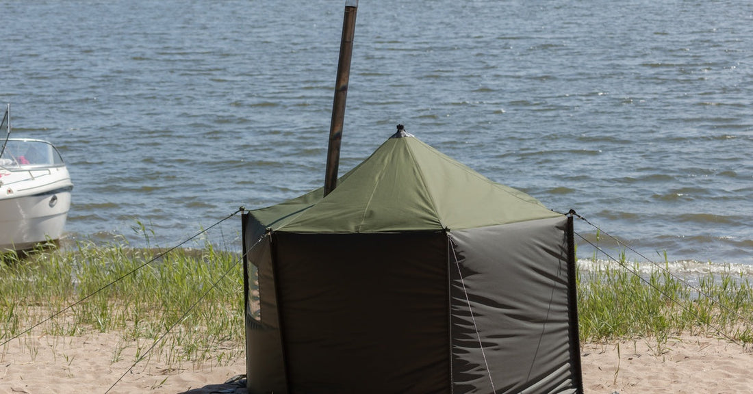 A single sauna tent is set up in a sandy, grassy area by a body of water. A boater speeds by in the background.