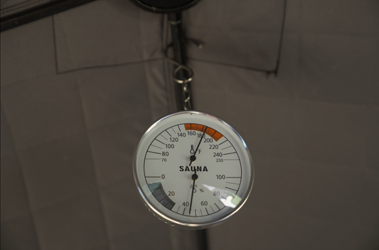 An analog sauna thermometer hygrometer with a white dial, black markings, and a glass front, hanging from a chain.